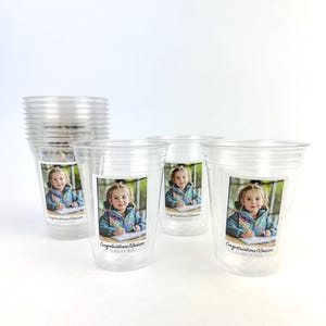 May include: Clear plastic cups with a printed image of a young girl writing. The image is on a white background. The text "Congratulations Madison CLASS OF 2026" is printed below the image. A stack of cups is visible.