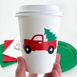 May include: A white paper coffee cup with a white lid. The cup features a red truck graphic carrying a green Christmas tree. Black wheels complete the truck design. The cup is held in a hand.
