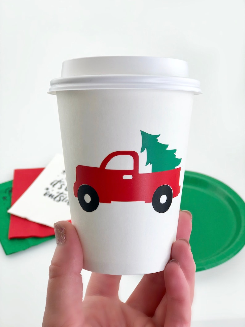 May include: A white paper coffee cup with a white lid. The cup features a red truck graphic carrying a green Christmas tree. Black wheels complete the truck design. The cup is held in a hand.