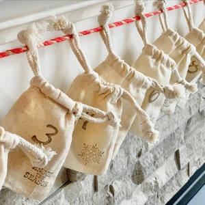 Puede incluir: Calendario de Adviento con peque&ntilde;as bolsas beige con cord&oacute;n colgando de una cuerda roja y blanca. Cada bolsa est&aacute; numerada con una fuente marr&oacute;n, y algunas presentan un dise&ntilde;o de copo de nieve. El texto "ENJOY THE SEASON" est&aacute; impreso en una de las bolsas.