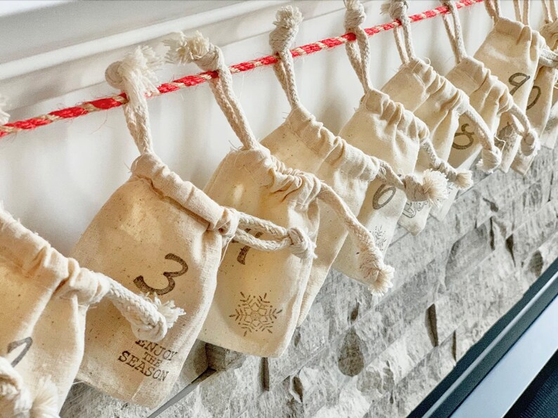 Puede incluir: Calendario de Adviento con peque&ntilde;as bolsas beige con cord&oacute;n colgando de una cuerda roja y blanca. Cada bolsa est&aacute; numerada con una fuente marr&oacute;n, y algunas presentan un dise&ntilde;o de copo de nieve. El texto "ENJOY THE SEASON" est&aacute; impreso en una de las bolsas.