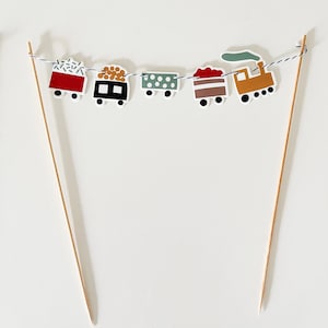 Op de afbeelding: Een kleurrijke trein caketopper met vijf wagons, waaronder een rode wagon met witte sneeuwvlokken, een zwarte wagon met bruine boomstammen, een groene wagon met witte stippen, een rood-wit gestreepte wagon en een gele wagon met groene rook. De wagons zijn aan elkaar geregen met een blauw-wit touwtje en bevestigd aan twee houten spiesjes.