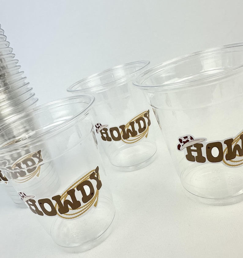 Puede incluir: Vasos de pl&aacute;stico transparente con un gr&aacute;fico marr&oacute;n "HOWDY" con un sombrero de vaquero y un dise&ntilde;o de cuerda. Los vasos est&aacute;n apilados y dispuestos sobre una superficie blanca, adecuados para un evento o fiesta tem&aacute;tica del oeste.