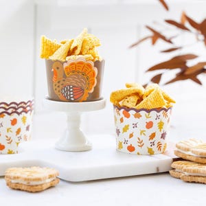 May include: Thanksgiving-themed snack cups filled with chips, alongside cookies. One cup features a turkey design, while others have fall leaf and pumpkin patterns. The cups sit on a white surface with a decorative background.