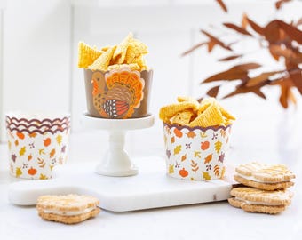 Op de afbeelding: Thanksgiving-thema snackbekers gevuld met chips, naast koekjes. Eén beker heeft een kalkoenontwerp, terwijl andere herfstblad- en pompoenpatronen hebben. De bekers staan op een wit oppervlak met een decoratieve achtergrond.