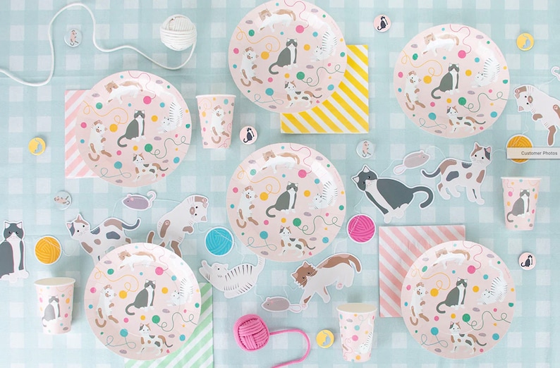 May include: A set of pink and white cat-themed party supplies, including plates, cups, napkins, and decorations. The plates and cups feature a pink background with colorful cats, yarn balls, and mice. The napkins have a pink and white striped pattern. The decorations include cat-shaped cutouts and yarn balls.