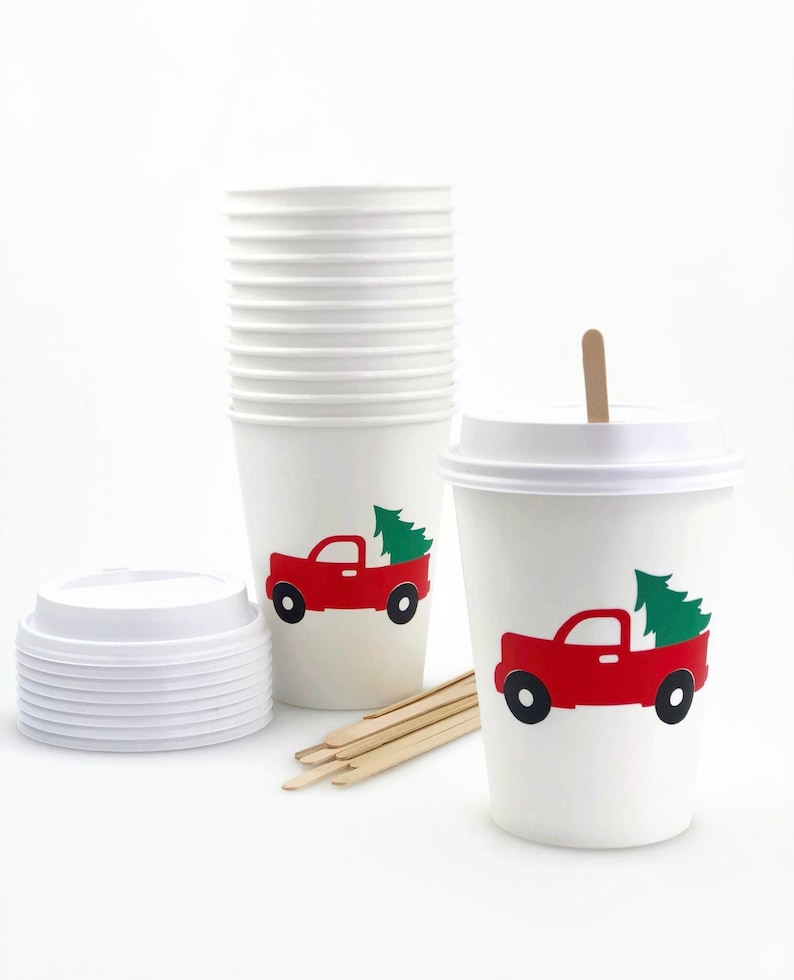 May include: White paper coffee cups with lids and stir sticks. Each cup features a red truck carrying a green Christmas tree. The cups are stacked and ready for use, perfect for holiday beverages.