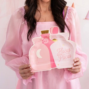 May include: Pink paper party plates with a design of three potion bottles and the text "Hocus All Your Pocus".