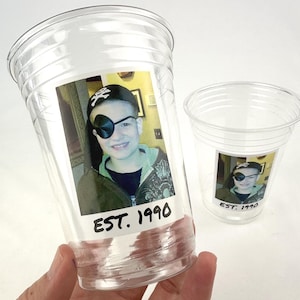 May include: Clear plastic cups with a printed photo of a person wearing a pirate hat and eye patch. The photo has the text "EST. 1990" printed below the image. One cup is held in a hand, and another cup is in the background.