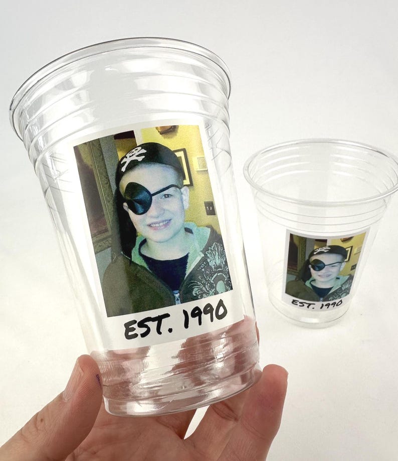 May include: Clear plastic cups with a printed photo of a person wearing a pirate hat and eye patch. The photo has the text "EST. 1990" printed below the image. One cup is held in a hand, and another cup is in the background.