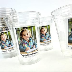 May include: Clear plastic cups with a printed label featuring a young girl holding a pencil and writing. The label reads "Congratulations Madison CLASS OF 2025". The cups are stacked and arranged on a white surface.