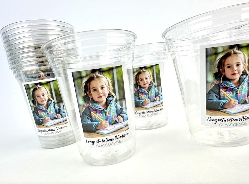 May include: Clear plastic cups with a printed label featuring a young girl holding a pencil and writing. The label reads "Congratulations Madison CLASS OF 2025". The cups are stacked and arranged on a white surface.
