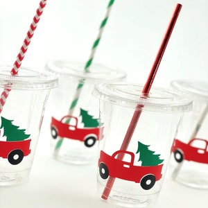 K&ouml;nnte beinhalten: Vier durchsichtige Plastikbecher mit roten und gr&uuml;nen Weihnachts-LKW-Designs und rot-wei&szlig; gestreiften Strohhalmen.