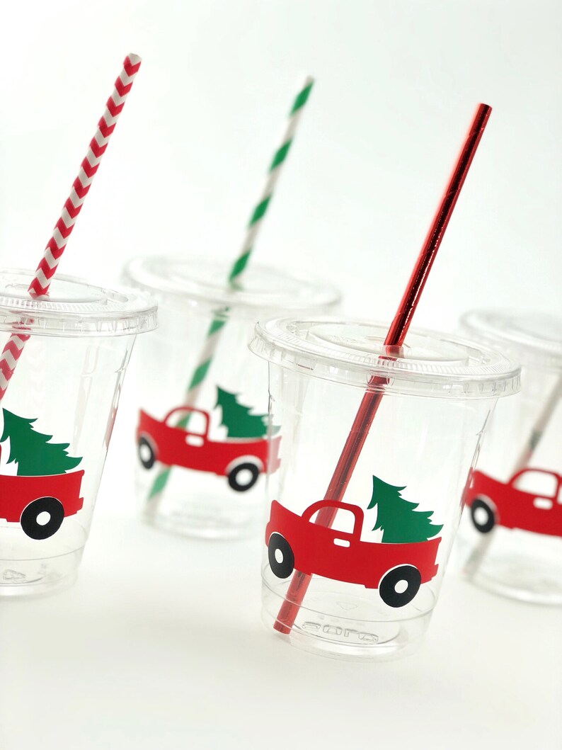 K&ouml;nnte beinhalten: Vier durchsichtige Plastikbecher mit roten und gr&uuml;nen Weihnachts-LKW-Designs und rot-wei&szlig; gestreiften Strohhalmen.