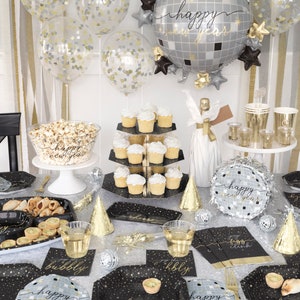 May include: Black and gold New Year's Eve party supplies with a disco ball theme. The set includes plates, napkins, cups, a cake stand, a popcorn bowl, and a balloon. The balloons are clear with gold confetti and a large silver disco ball balloon with the text "happy new year".