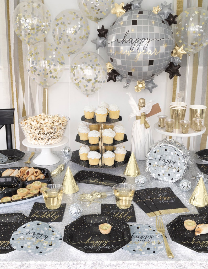 May include: Black and gold New Year's Eve party supplies with a disco ball theme. The set includes plates, napkins, cups, a cake stand, a popcorn bowl, and a balloon. The balloons are clear with gold confetti and a large silver disco ball balloon with the text "happy new year".