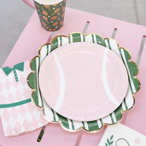 Puede incluir: Un plato de papel rosa con un dise&ntilde;o de pelota de tenis se encuentra sobre un plato festoneado verde y blanco con borde dorado. Tambi&eacute;n se ven una taza y una servilleta a juego con motivos de tenis, todo sobre una mesa rosa.