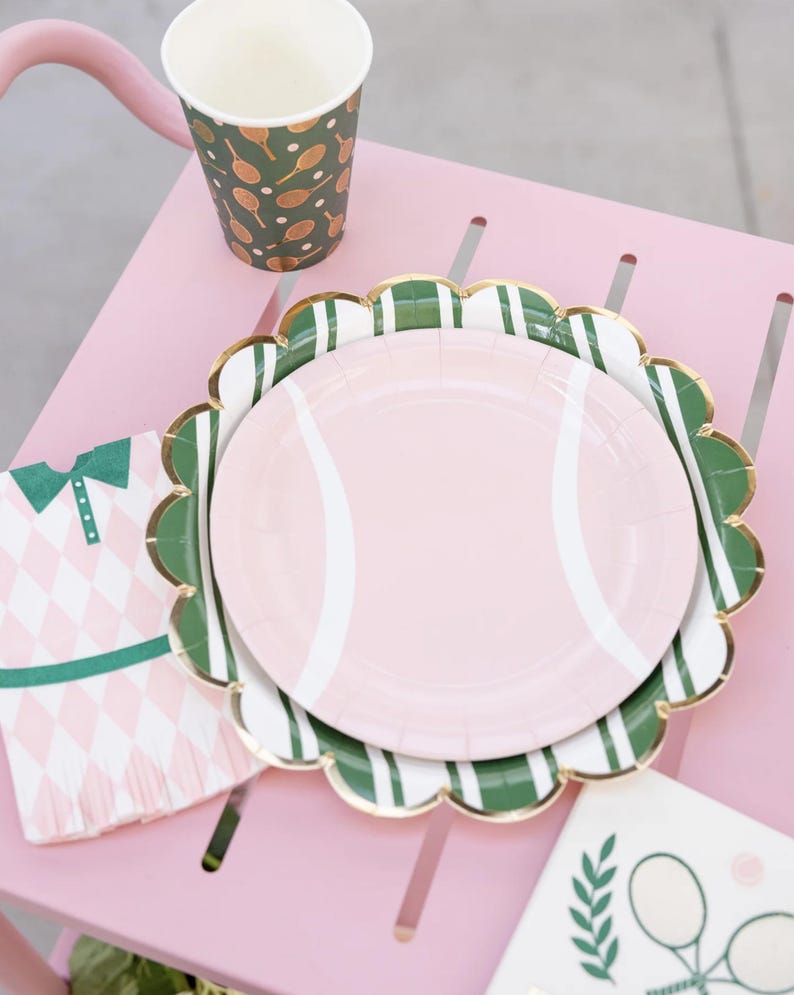 Puede incluir: Un plato de papel rosa con un dise&ntilde;o de pelota de tenis se encuentra sobre un plato festoneado verde y blanco con borde dorado. Tambi&eacute;n se ven una taza y una servilleta a juego con motivos de tenis, todo sobre una mesa rosa.