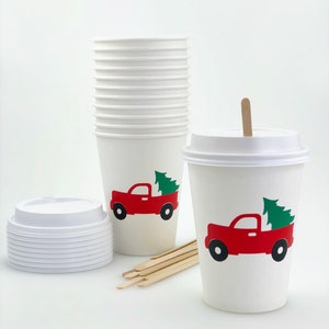 May include: White paper coffee cups with lids, stir sticks, and a stack of cups. Each cup features a red truck carrying a green Christmas tree. The cups are designed for hot beverages.
