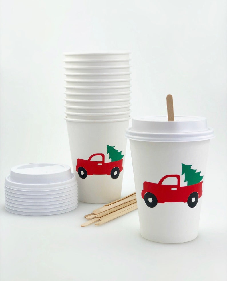May include: White paper coffee cups with lids, stir sticks, and a stack of cups. Each cup features a red truck carrying a green Christmas tree. The cups are designed for hot beverages.