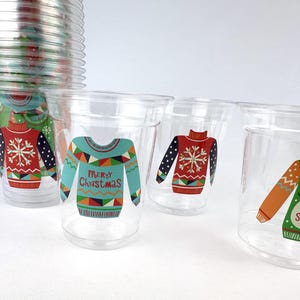 May include: Clear plastic cups with festive Christmas sweater designs. The cups feature various sweater patterns in red, blue, and green, with snowflake and holiday text details. A stack of similar cups is visible in the background.
