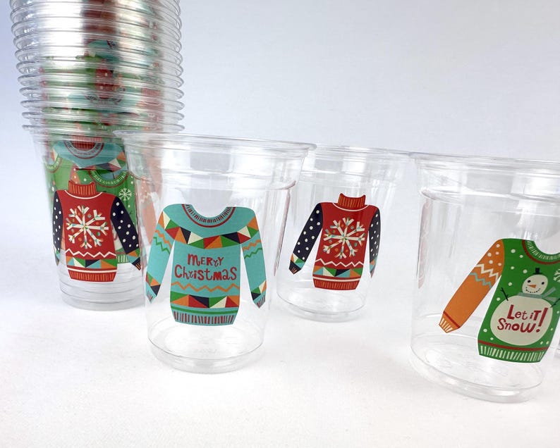 May include: Clear plastic cups with festive Christmas sweater designs. The cups feature various sweater patterns in red, blue, and green, with snowflake and holiday text details. A stack of similar cups is visible in the background.