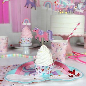 May include: A pink and purple unicorn cupcake topper with a rainbow on top. The cupcake is on a white cupcake liner with pink and gold hearts. The cupcake is on a blue and pink paper plate with a unicorn design. There are pink and white striped straws and pink and white cups with unicorn designs in the background.