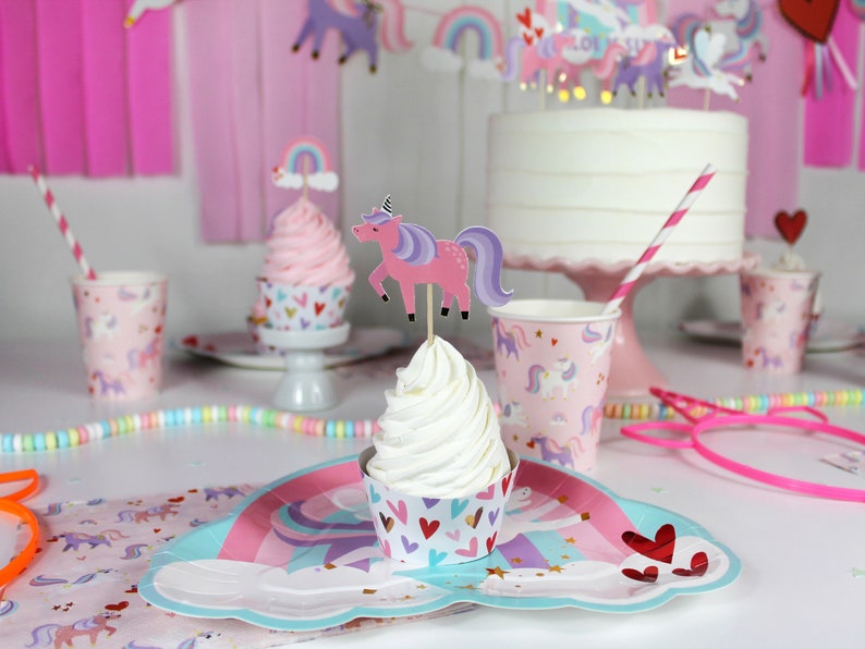 May include: A pink and purple unicorn cupcake topper with a rainbow on top. The cupcake is on a white cupcake liner with pink and gold hearts. The cupcake is on a blue and pink paper plate with a unicorn design. There are pink and white striped straws and pink and white cups with unicorn designs in the background.