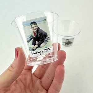 Puede incluir: Vasos de chupito de plástico transparente con una foto de una persona en la playa. La foto está etiquetada como "Vintage 2000". Un vaso se sostiene en la mano y otro está en el fondo.