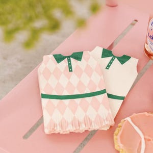 May include: Two paper napkins shaped like golf shirts. One napkin has a pink and white diamond pattern, green collar and trim. The other is white with a green collar and trim. The napkins are on a pink surface.