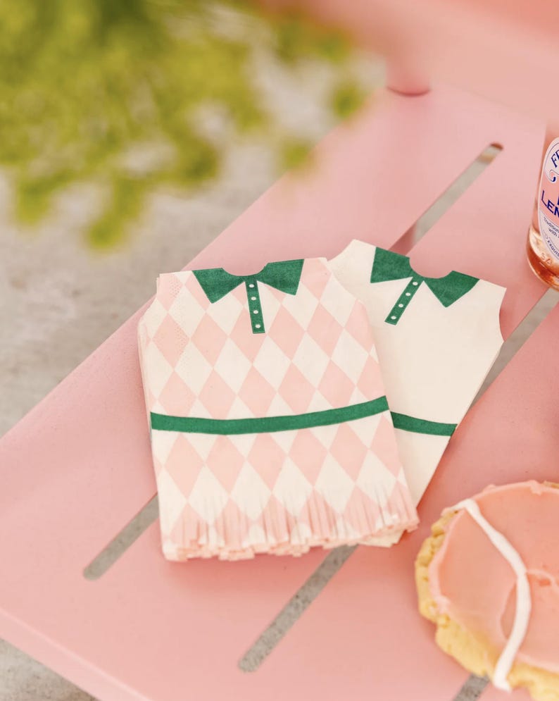 Puede incluir: Dos servilletas de papel con forma de camisas de golf. Una servilleta tiene un patr&oacute;n de diamantes rosas y blancos, cuello y ribete verdes. La otra es blanca con cuello y ribete verdes. Las servilletas est&aacute;n sobre una superficie rosa.