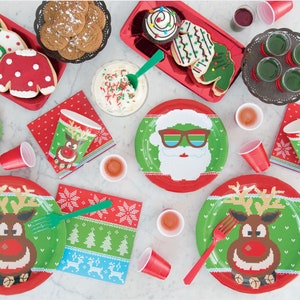 Pu&ograve; includere: Una tavola imbandita per una festa di Natale con piatti, bicchieri e tovaglioli di carta rossi e verdi con un renna e Babbo Natale in un design di maglione festivo. Ci sono anche biscotti e cupcake decorati con luci natalizie e una ghirlanda.