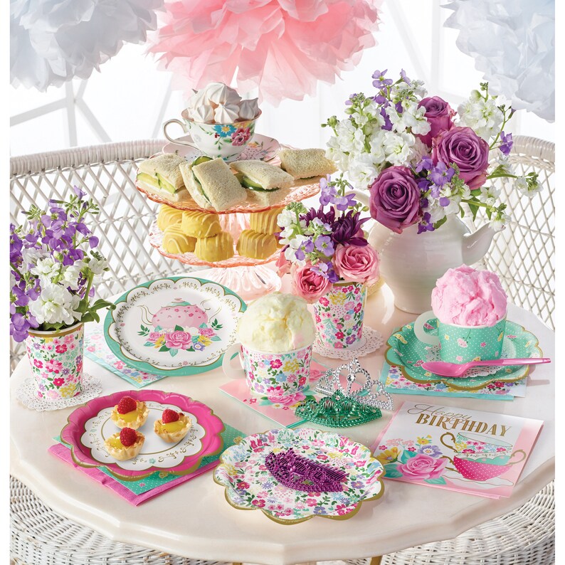May include: A pink and green tea party table setting with floral patterned paper plates, cups, napkins, and a tiered cake stand with sandwiches and cupcakes. There is a pink and green teacup with a pink cotton candy dessert, a green crown, and a card that says "Happy Birthday".