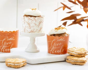 Op de afbeelding: Een stilleven met cupcakes met witte glazuur en strooisel in decoratieve papieren bekers. De bekers zijn oranje met de woorden "Give Thanks" in witte letters. Er zijn ook twee sandwichkoekjes aanwezig.