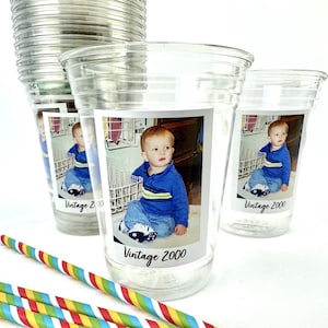 Puede incluir: Vasos de pl&aacute;stico transparente con una foto de un ni&ntilde;o peque&ntilde;o y el texto "Vintage 2000". El ni&ntilde;o lleva una camisa azul y vaqueros. Hay pajitas de colores a rayas en primer plano.