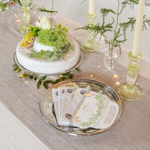 May include: A white cake with green foliage and a white rose on top, sitting on a silver cake stand. A silver platter with a stack of white napkins with a floral design and the text "Thank You" is on a linen tablecloth. There are two green glass candle holders with white candles on the table.