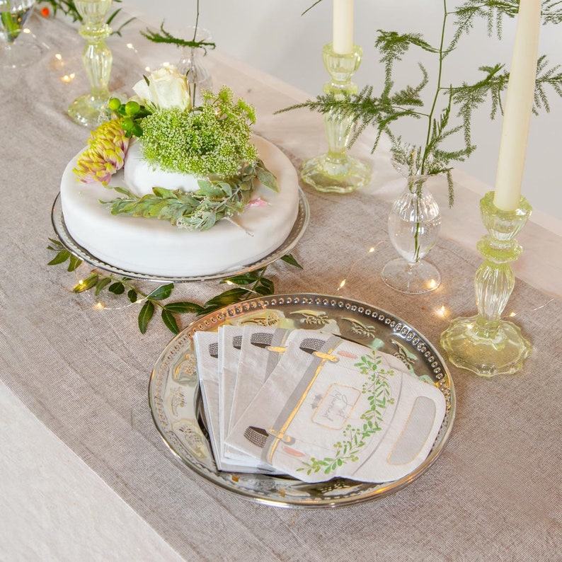 May include: A white cake with green foliage and a white rose on top, sitting on a silver cake stand. A silver platter with a stack of white napkins with a floral design and the text "Thank You" is on a linen tablecloth. There are two green glass candle holders with white candles on the table.