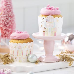 May include: Two pink frosted cupcakes decorated with nutcracker themed cupcake liners. The cupcakes are on a pink cake stand and a white surface with gold glitter ornaments.