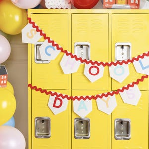Może przedstawiać: Żółty zestaw szafek ozdobiony czerwono-białym banerem z napisem "SCHOOL DAYS" w kolorowych literach. Po lewej stronie zdjęcia widoczne są balony w różnych kolorach, w tym żółtym, różowym i czerwonym. Miniaturowy budynek szkoły stoi na balonach.
