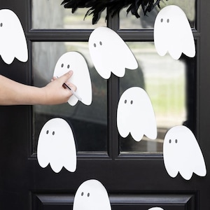 Op de afbeelding: Witte spookvormige uitsparingen met zwarte stipogen zijn bevestigd aan een zwarte deur. Een hand tekent op een van de geesten met een pen. Een zwarte verenkrans bevindt zich boven de deur en creëert een Halloween-thema.