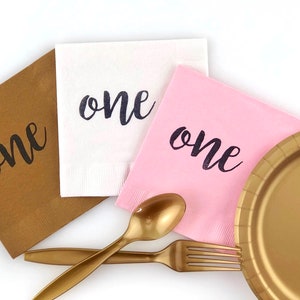 May include: Three paper napkins with the word "one" printed on them in black script. The napkins are brown, white, and pink. There is a gold plastic plate, a gold plastic fork, and a gold plastic spoon on a white surface.