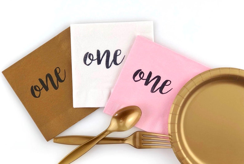 May include: Three paper napkins with the word "one" printed on them in black script. The napkins are brown, white, and pink. There is a gold plastic plate, a gold plastic fork, and a gold plastic spoon on a white surface.
