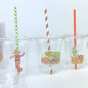 May include: Three clear plastic cups with lids and straws. Each cup has a different fall-themed sticker. One cup has a scarecrow, one has a tractor, and one has a corn stalk and pumpkins.