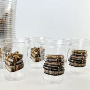 Könnte beinhalten: Klare Plastikbecher, verziert mit Aufklebern von gestapelten Vintage-Büchern, die mit goldenen Bändern gebunden sind. Die Becher sind in einer Gruppe angeordnet, mit einem Stapel zusätzlicher Becher im Hintergrund. Die Bücher haben ein detailliertes, gealtertes Aussehen.