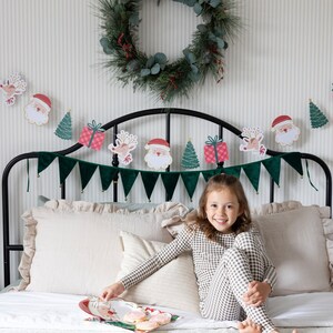 May include: A young girl wearing a brown and white checked pyjama set sits on a bed with a white and green patterned bedspread. A Christmas garland with Santa and reindeer characters hangs above the bed. A green wreath with red berries hangs on the wall behind the bed. The girl is holding a plate with cookies on it. The text "COORDINATING PARTY SUPPLIES AVAILABLE" is printed on the wall.