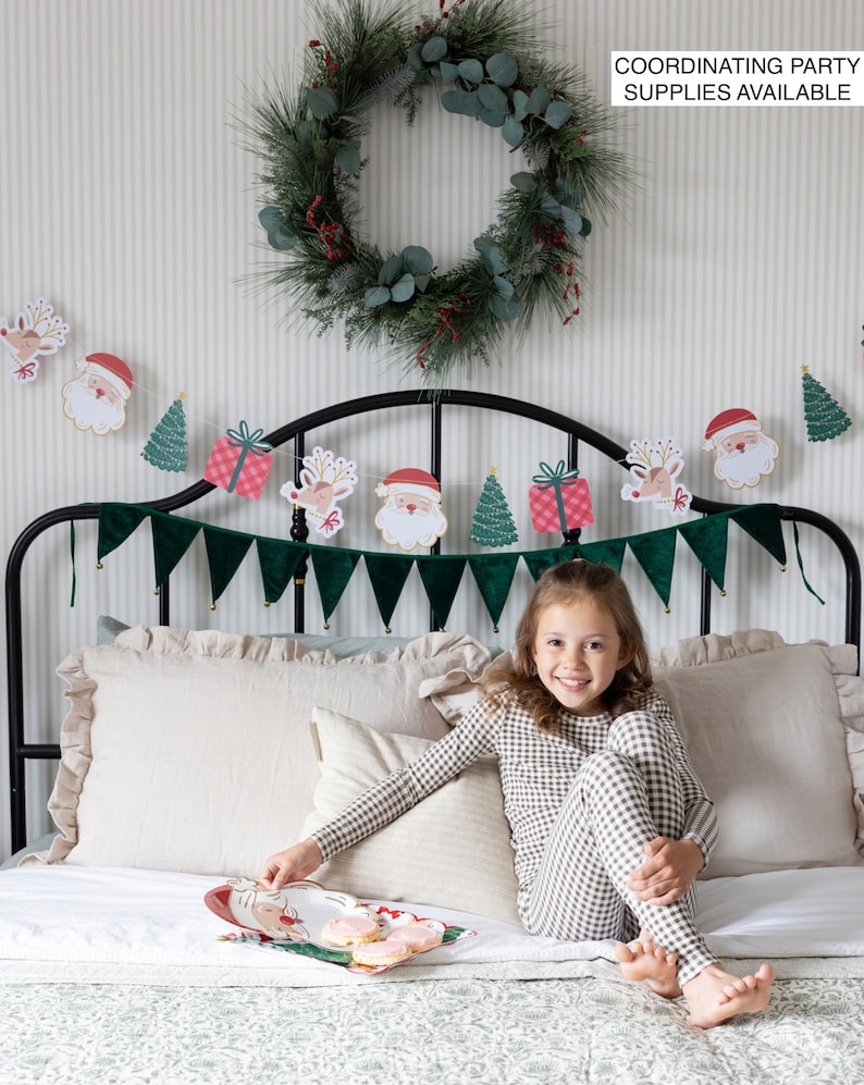May include: A young girl wearing a brown and white checked pyjama set sits on a bed with a white and green patterned bedspread. A Christmas garland with Santa and reindeer characters hangs above the bed. A green wreath with red berries hangs on the wall behind the bed. The girl is holding a plate with cookies on it. The text "COORDINATING PARTY SUPPLIES AVAILABLE" is printed on the wall.