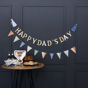 K&ouml;nnte beinhalten: Ein Happy Dad's Day Banner mit blauen und goldenen Wimpeln und einem goldenen Happy Dad's Day Banner an einer dunkelgrauen Wand. Im Vordergrund steht ein Tisch mit einer Troph&auml;e, Snacks und Getr&auml;nken.
