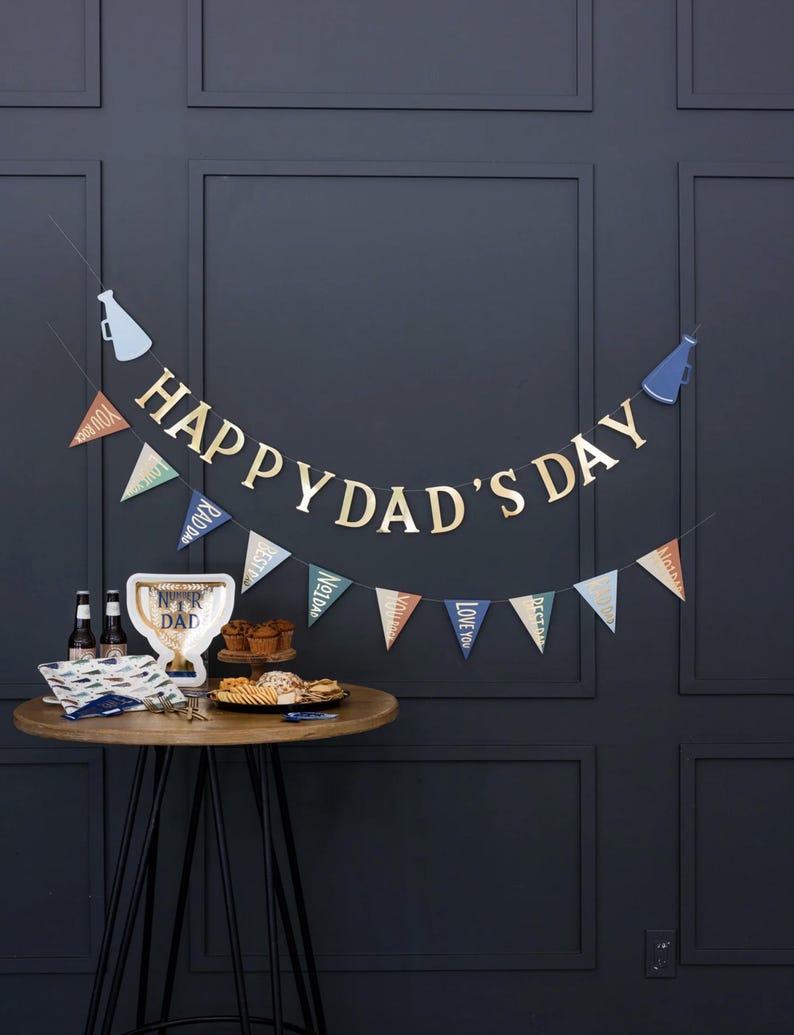 K&ouml;nnte beinhalten: Ein Happy Dad's Day Banner mit blauen und goldenen Wimpeln und einem goldenen Happy Dad's Day Banner an einer dunkelgrauen Wand. Im Vordergrund steht ein Tisch mit einer Troph&auml;e, Snacks und Getr&auml;nken.