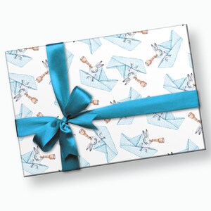 Puede incluir: Un regalo envuelto con papel blanco con un patr&oacute;n de barcos de papel azules y conejos mirando a trav&eacute;s de telescopios. Una cinta de color turquesa est&aacute; atada alrededor del regalo.