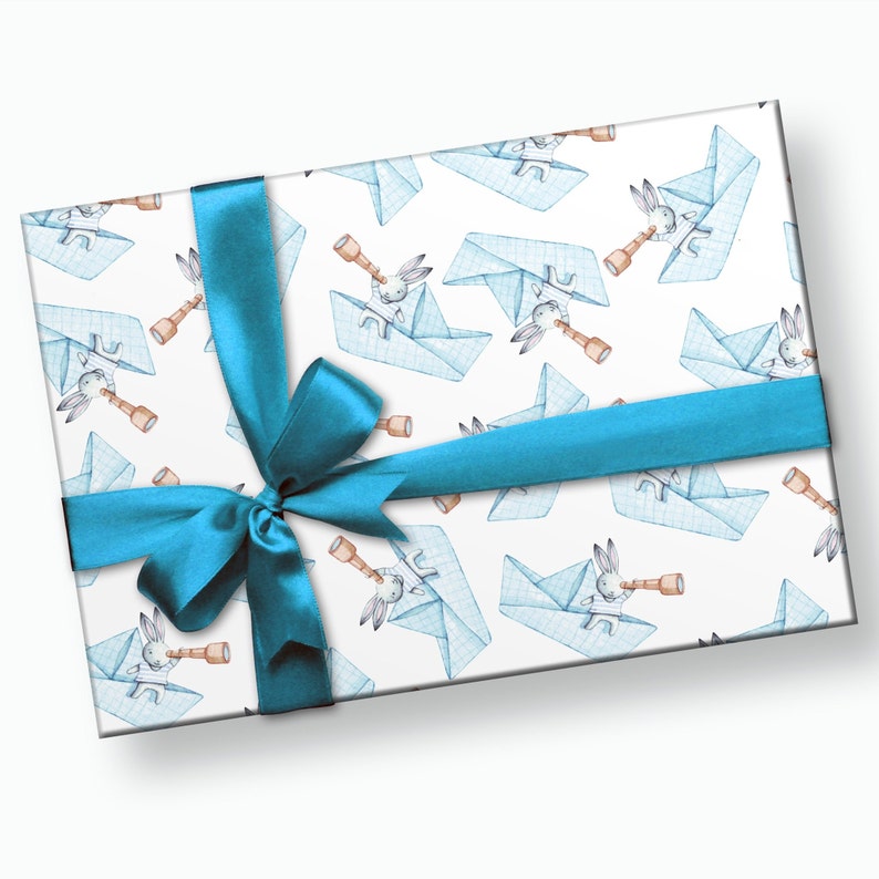 Puede incluir: Un regalo envuelto con papel blanco con un patr&oacute;n de barcos de papel azules y conejos mirando a trav&eacute;s de telescopios. Una cinta de color turquesa est&aacute; atada alrededor del regalo.